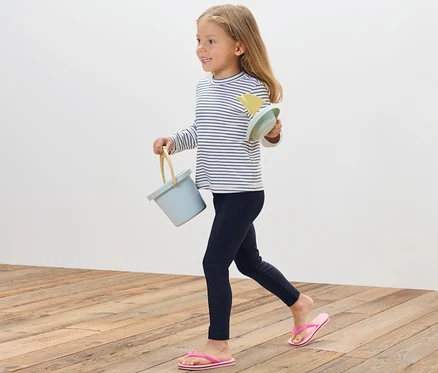Ein Mädchen geht auf einem Holzboden und trägt einen blauen Eimer und Sandspielzeug. Sie trägt eine Kinder-Badeleggings mit UV-Schutzfaktor 80.
