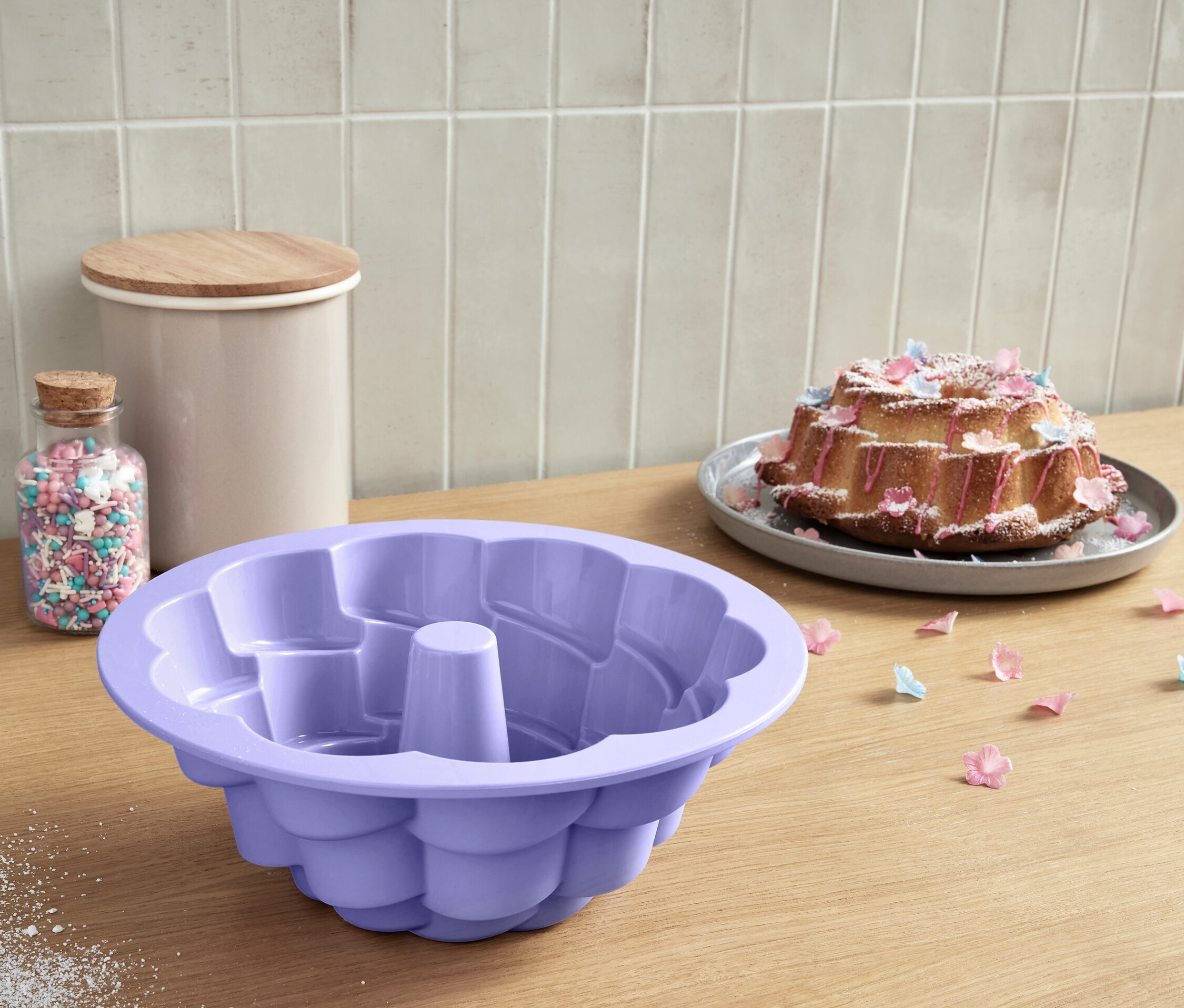 Eine lila Gugelhupfform Blüte liegt auf einem Holztisch neben einem Glas mit Streuseln.