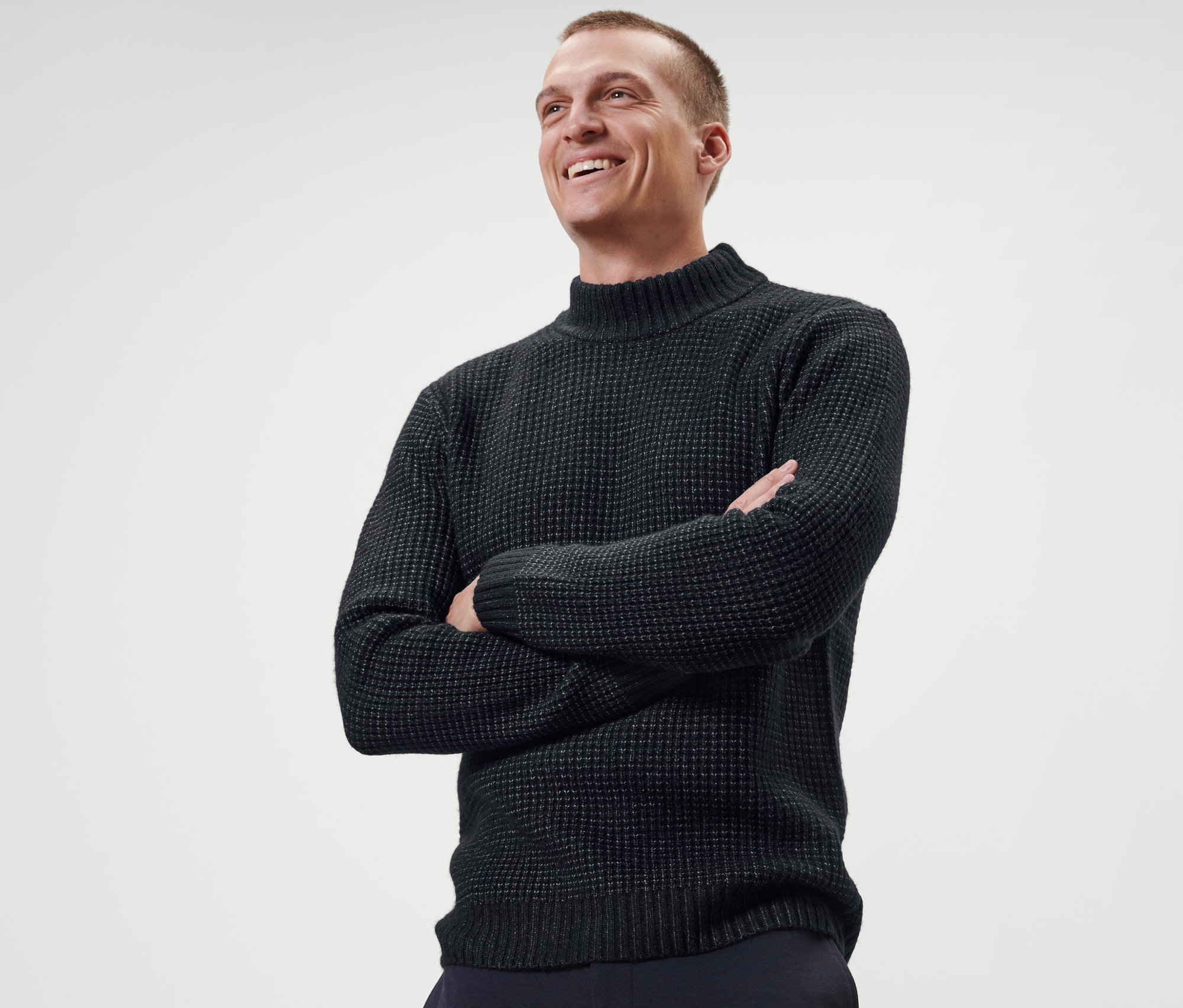 Lächelnder Mann posiert mit verschränkten Armen in einem Strickpullover.