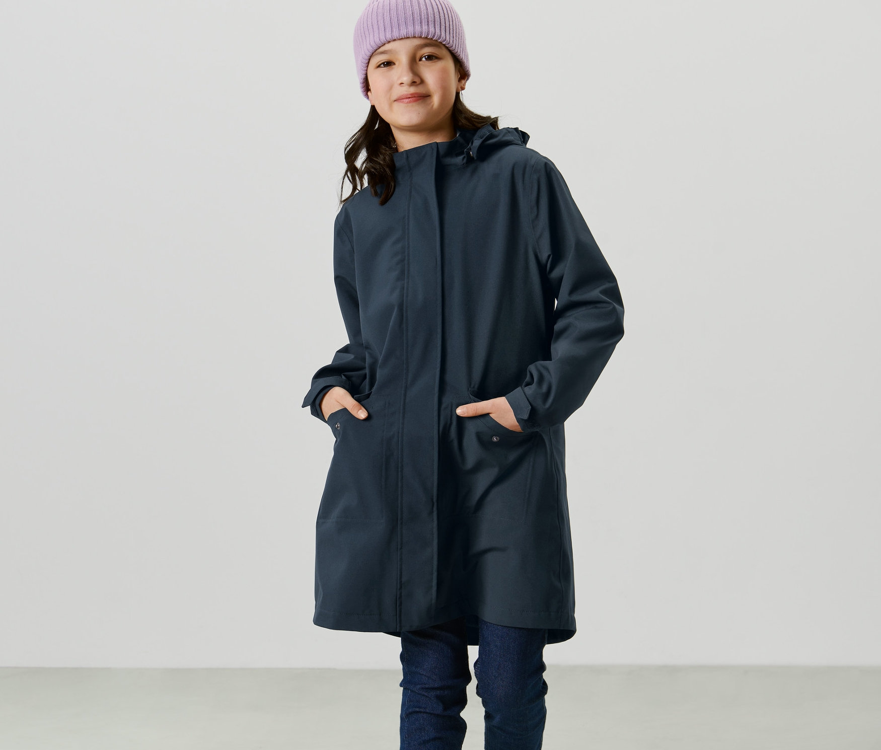 Mädchen mit lila Mütze trägt einen Kinder-Regenparka und steht mit Händen in den Taschen.