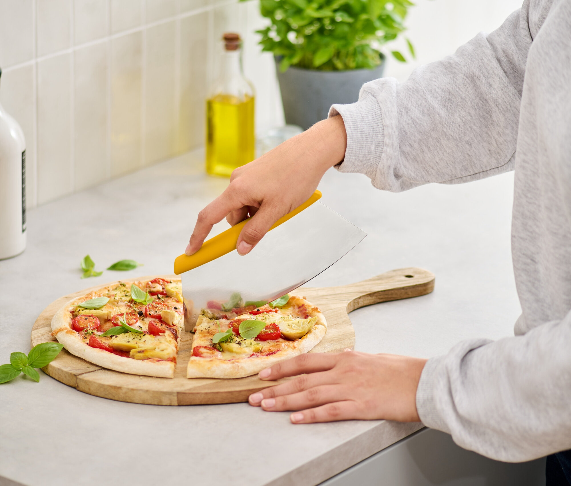 Frau schneidet Pizza mit Tomaten und Basilikum mit einem gelben Pizzaschneider auf einem Holzbrett.
