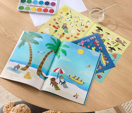 Ein geöffnetes Mein Stickerbuch »Am Meer« mit Strandillustrationen, Farben zum Malen und Stickerbögen auf einem Holztisch.