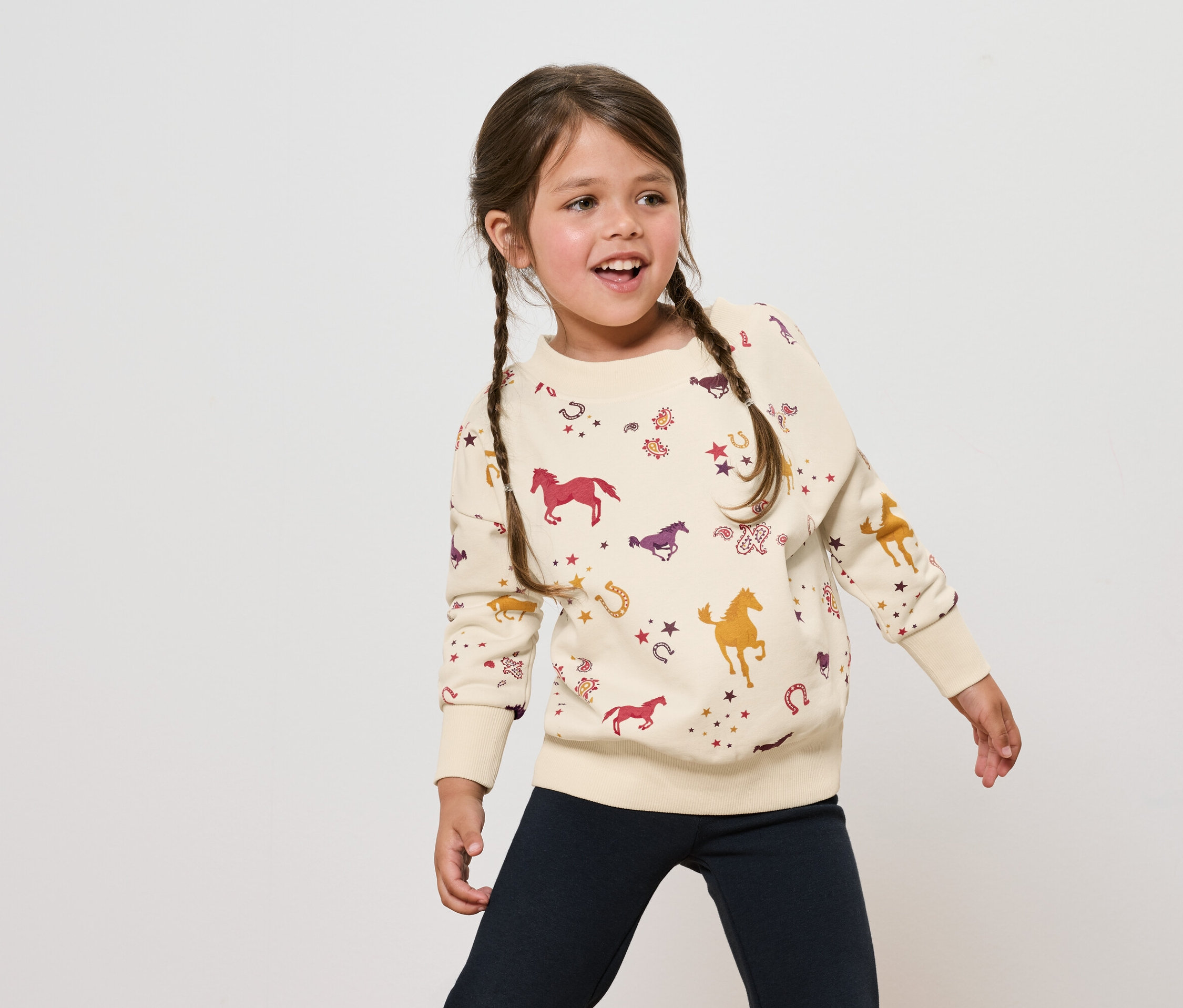 Mädchen mit braunen Haaren in geflochtenen Zöpfen trägt ein cremefarbenes Sweatshirt mit Pferdeaufdruck und marineblaue Hosen.