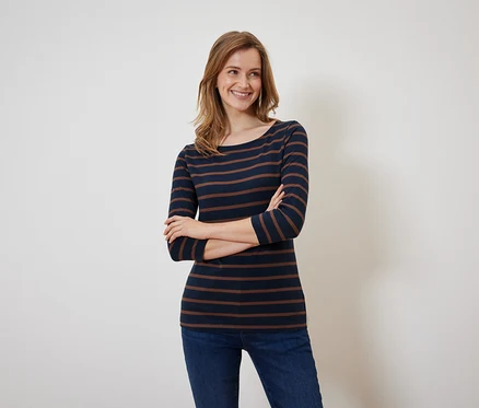 Eine lächelnde Frau posiert mit verschränkten Armen in einem marineblau-braun gestreiften Shirt und Jeans.