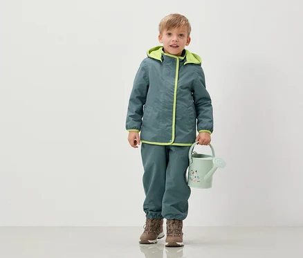 Ein Junge in Jacke und Hose hält eine hellgrüne Gießkanne.