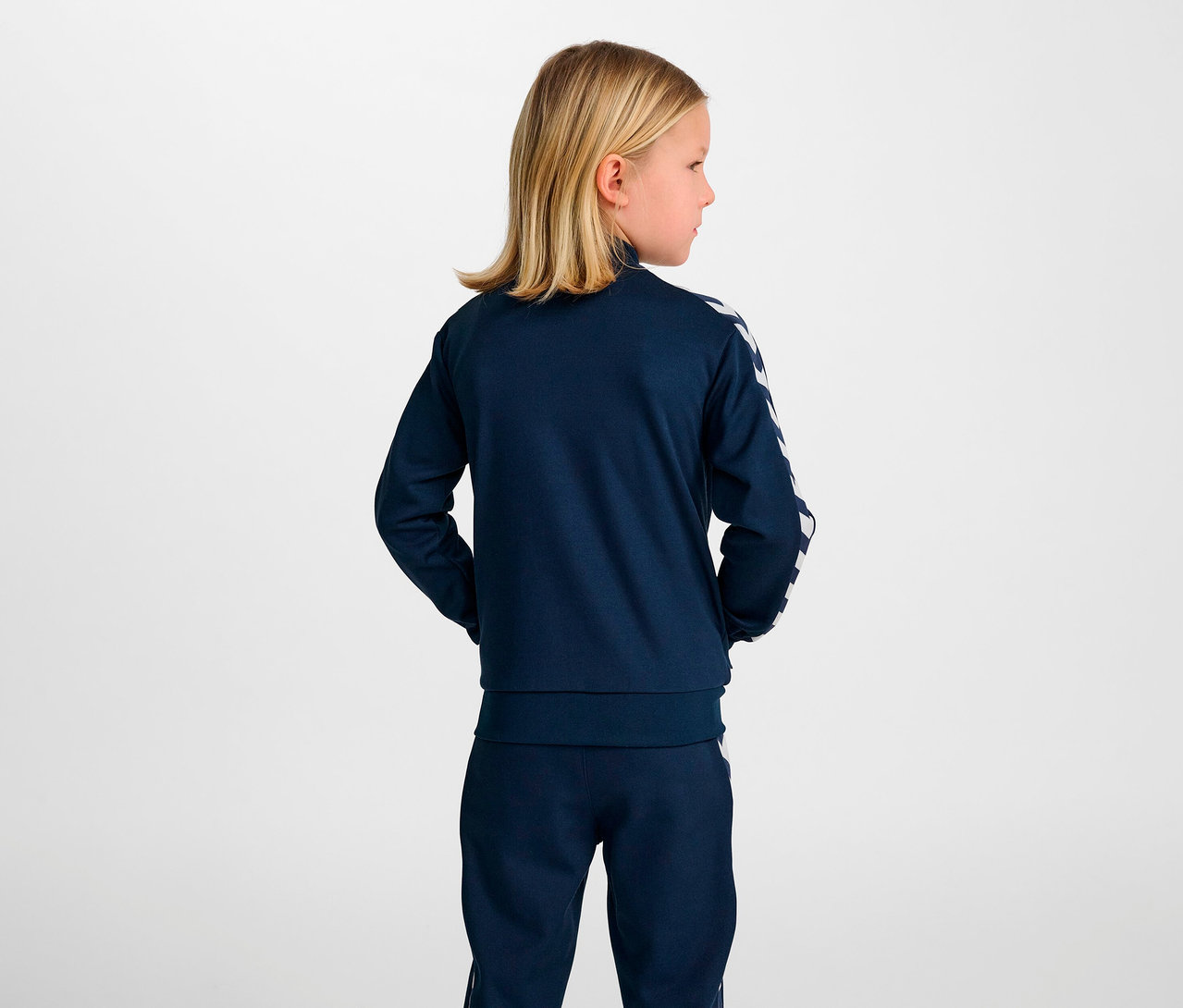 Blondes Mädchen steht mit dem Rücken zur Kamera und trägt eine blaue HUMMEL HMLJR Archive Reg Poly Zip Jacket.