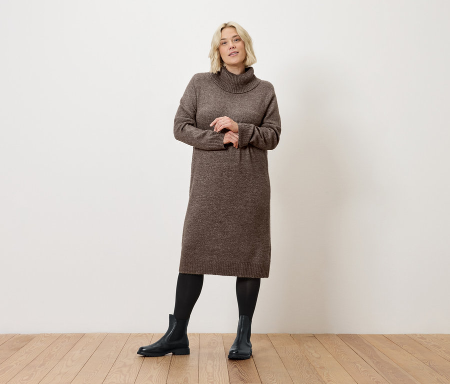 Blonde Frau posiert in einem braunen Rollkragenkleid, schwarzen Leggings und Stiefeln auf einem Holzboden vor einer weißen Wand.