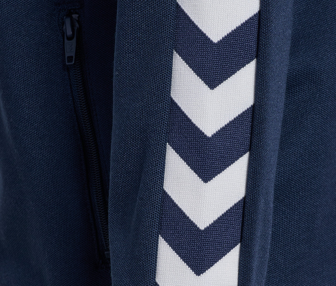 Detail einer blauen Hummel HMLJR Archive Reg Poly Zip Jacket mit weißen Winkelstreifen am Ärmel und Reißverschluss.