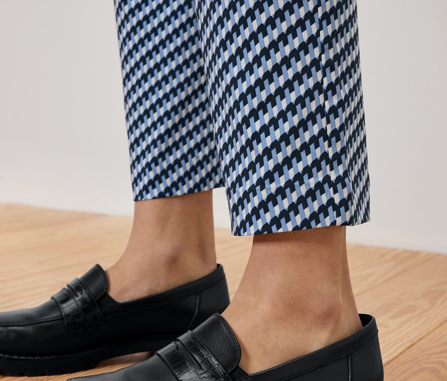 Beine einer Person in blau-weiss gemusterten Hosen und schwarzen Lederloafers auf einem Holzboden.