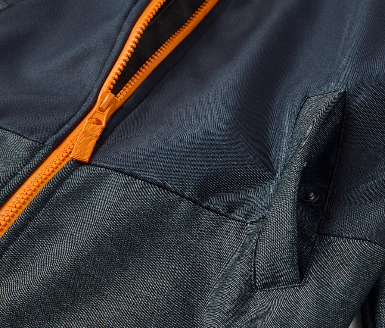 Detailaufnahme einer Kinder-Softshelljacke mit orangefarbenem Reißverschluss und Tasche.