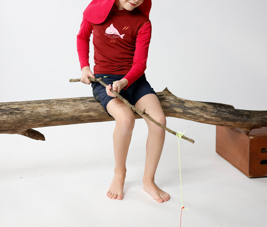 Ein Kind mit roter Mütze sitzt auf einem Baumstamm und angelt mit einem Stock.