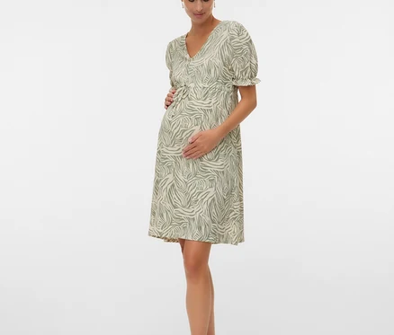 Schwangere Frau mit einem grün-weißen Kleid und Sandalen, die ihre Hände auf ihren Bauch legt.