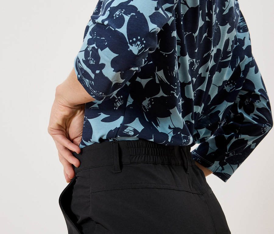 Eine Person mit einem hellblauen, blumengemusterten Oberteil und schwarzen Hosen steht vor einem weißen Hintergrund.