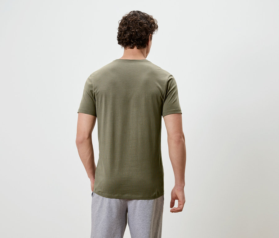 Ein Mann mit lockigem Haar steht mit dem Rücken zur Kamera und trägt ein olivgrünes T-Shirt und graue Shorts.
