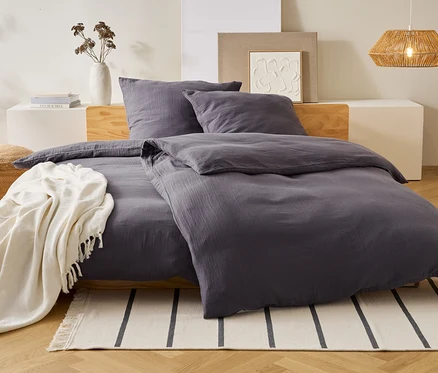 Graue Bettwäsche liegt auf einem Bett mit Kissen. Eine weiße Decke liegt über dem Bett. Eine Vase und eine Lampe stehen im Hintergrund.