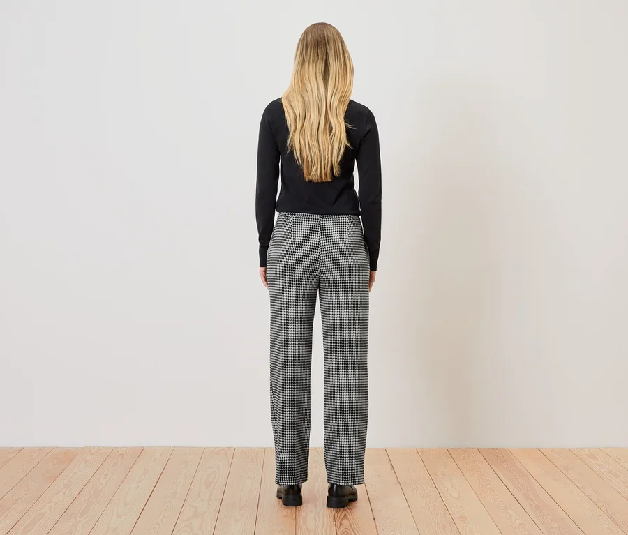 Eine Frau mit blonden Haaren steht mit dem Rücken zur Kamera und trägt ein schwarzes Oberteil und eine schwarz-weiß karierte Hose.