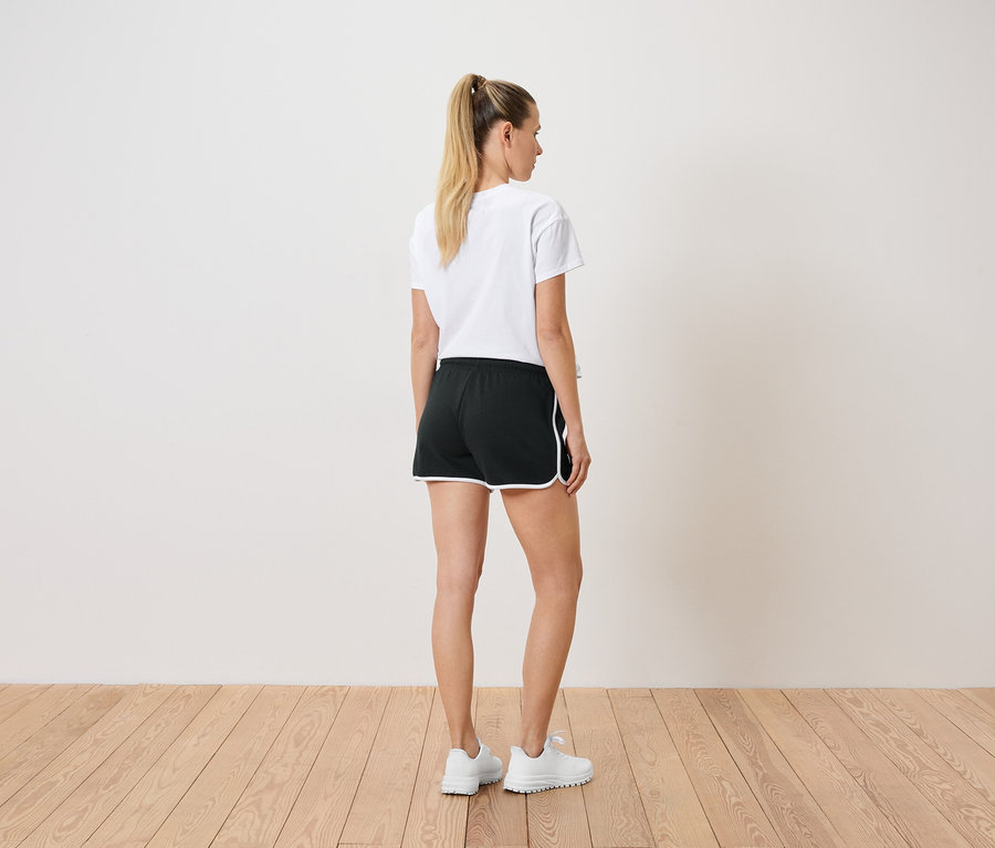 Frau mit Pferdeschwanz trägt ein weißes T-Shirt, schwarze Shorts mit weißem Rand und weiße Turnschuhe, von hinten gesehen.