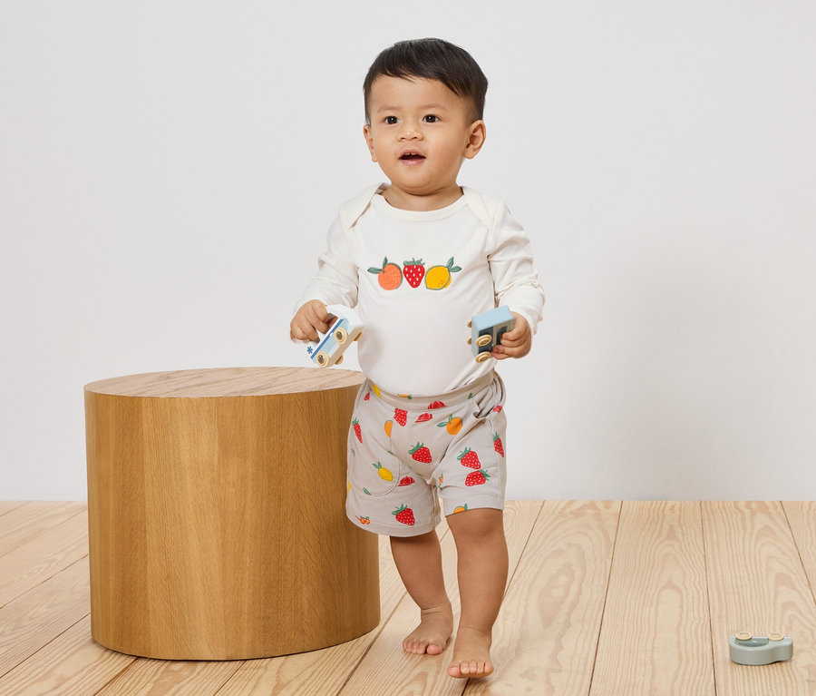 Ein Kleinkind steht barfuß auf einem Holzboden und hält zwei kleine Spielzeugautos in den Händen. Es trägt ein langärmeliges Oberteil mit Fruchtmotiven und kurze Hosen mit Erdbeermuster.