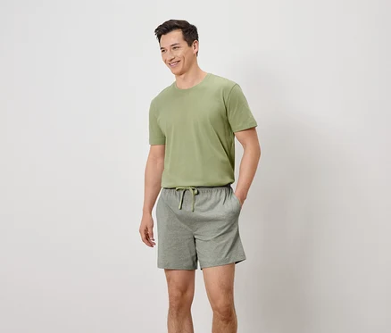 Mann mit olivgrünem T-Shirt und grauen Shorts mit Kordelzug, die Hände in den Hosentaschen, lächelnd.