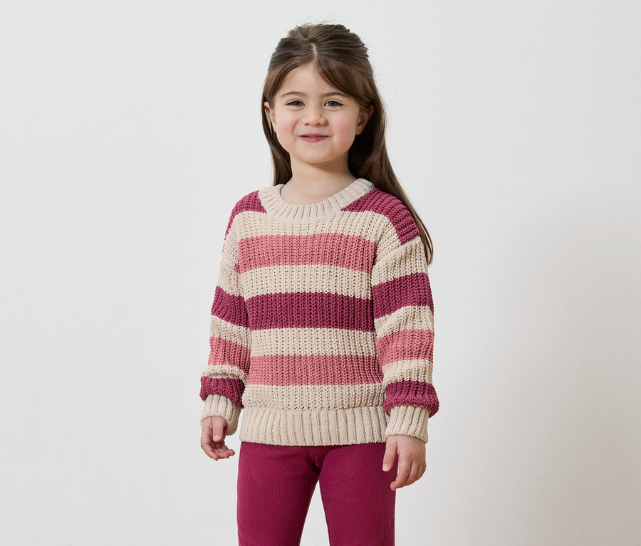 Mädchen mit braunen Haaren trägt einen gestreiften Pullover in Rosa, Beige und Burgunderrot und eine burgunderrote Hose vor einem weißen Hintergrund.