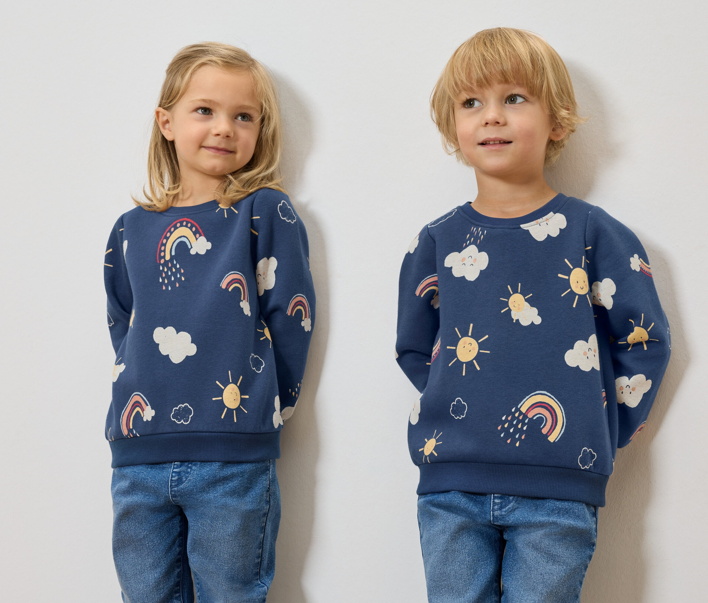 Zwei Kinder stehen nebeneinander und tragen dunkelblaue Kinder-Sweatshirts mit angerauter Innenseite und aufgedruckten Wolken, Sonnen und Regenbögen.