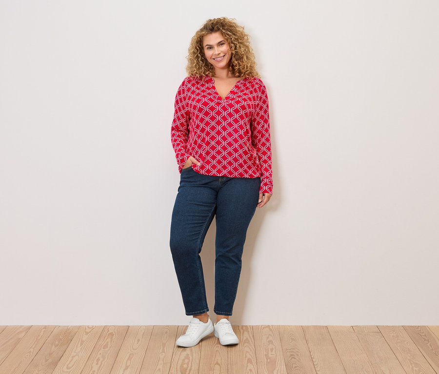 Frau mit lockigen blonden Haaren, roter Bluse mit weißem Muster, Jeans und weißen Turnschuhen steht vor einer Wand.