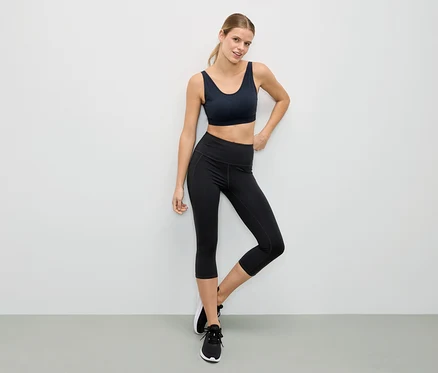 Frau posiert in einem schwarzen Sport-BH und schwarzen Leggings vor einer weißen Wand.