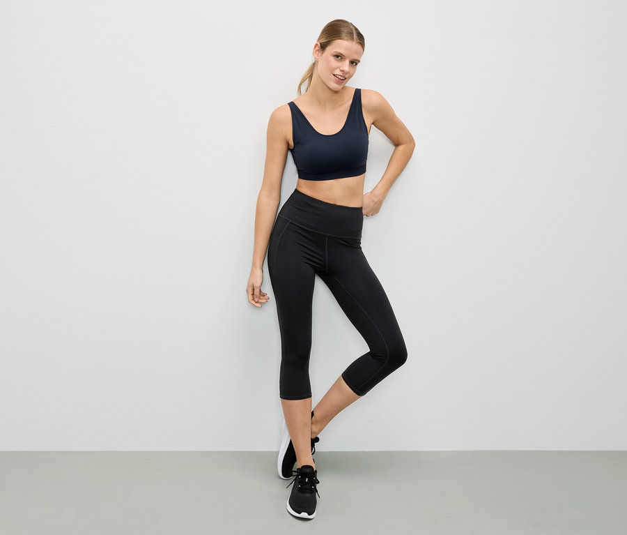 Frau posiert in einem schwarzen Sport-BH und schwarzen Leggings vor einer weißen Wand.