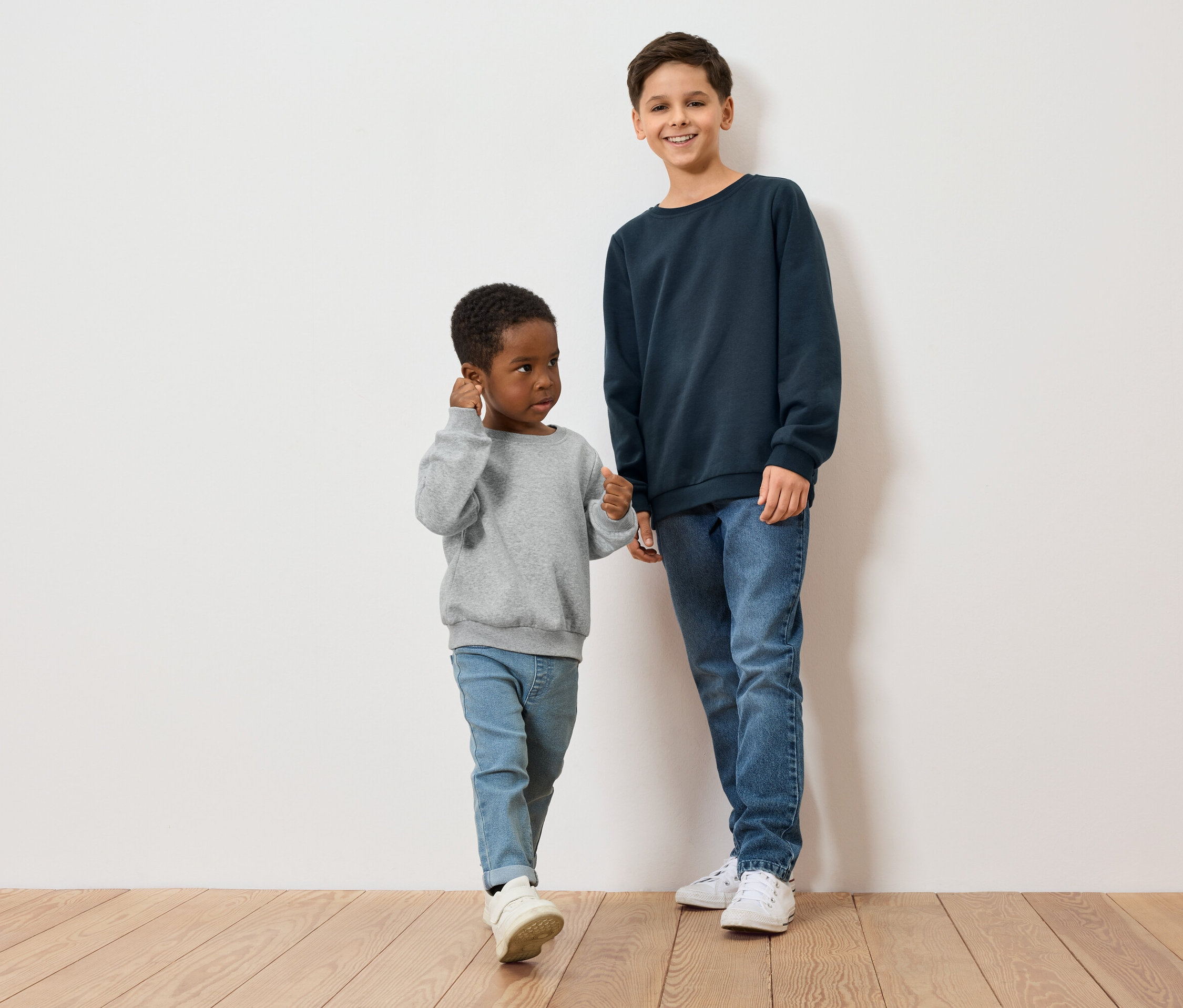 Zwei Jungen, einer mit dunkler Hautfarbe und grauem Pullover, der andere mit heller Hautfarbe und blauem Pullover, stehen auf einem Holzboden vor einer weißen Wand.