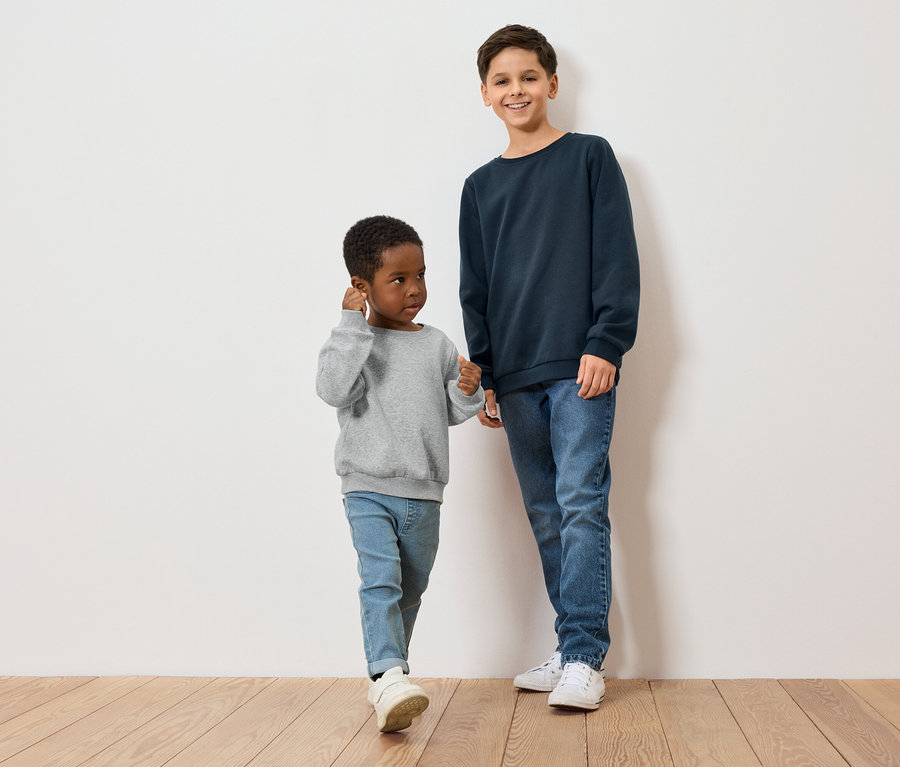 Zwei Jungen, einer mit dunkler Hautfarbe und grauem Pullover, der andere mit heller Hautfarbe und blauem Pullover, stehen auf einem Holzboden vor einer weißen Wand.