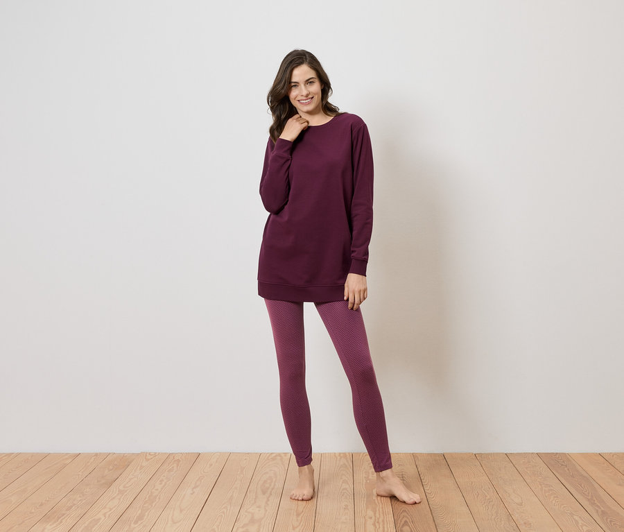 Eine Frau steht barfuß auf einem Holzboden vor einer weißen Wand und trägt ein weinrotes, langes Oberteil und Leggings.