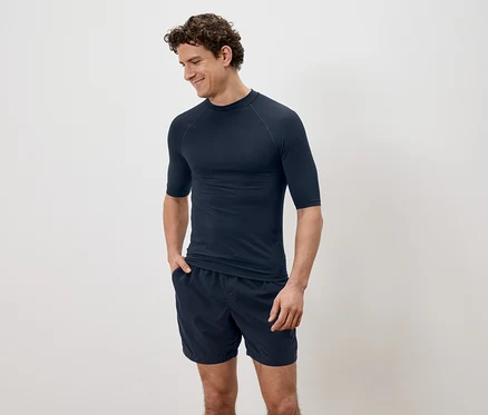 Mann mit lockigem Haar, der ein marineblaues, kurzärmeliges Hemd und Shorts vor einem schlichten Hintergrund trägt.
