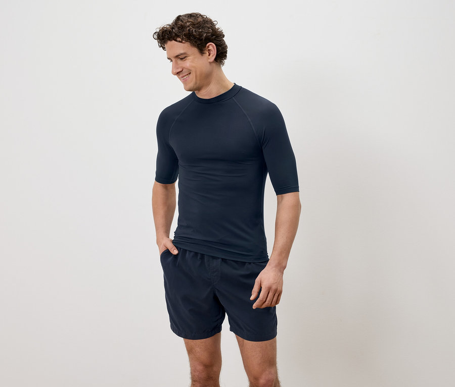 Mann mit lockigem Haar, der ein marineblaues, kurzärmeliges Hemd und Shorts vor einem schlichten Hintergrund trägt.