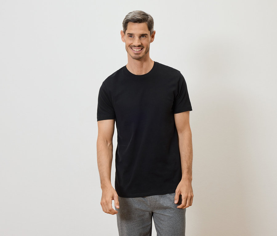Mann mit grau melierter Frisur lächelt in schwarzem T-Shirt und grauer Hose vor weißem Hintergrund.