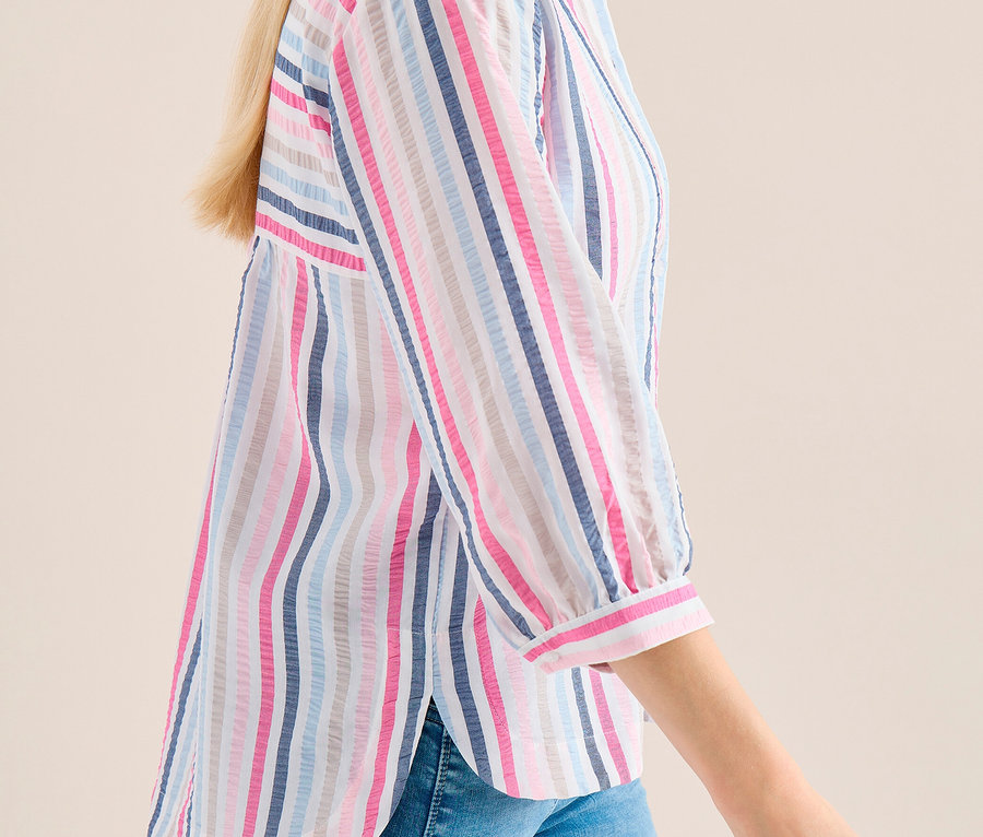Blonde Frau geht seitlich, trägt eine gestreifte CECIL Hemdbluse in Pink, Blau und Grau und blaue Jeans.