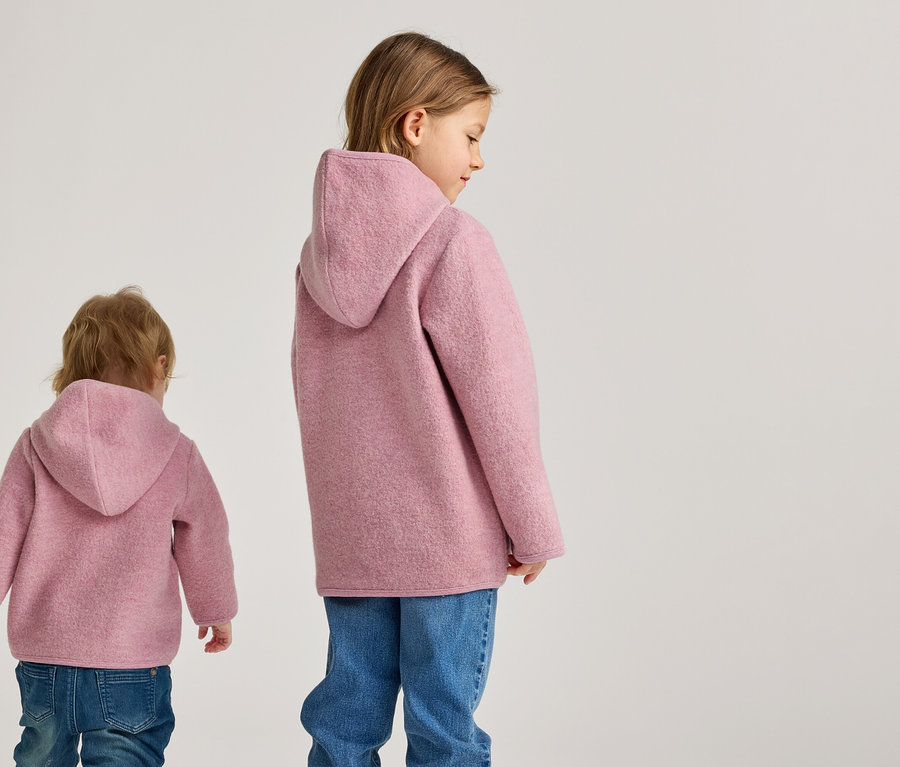 Zwei Kinder von hinten tragen rosa Wolljacken mit Kapuze und Jeans vor einem hellen Hintergrund.