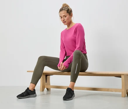 Frau sitzt auf einer Holzbank, trägt ein pinkes Langarmshirt, grüne Leggings und schwarze Turnschuhe.