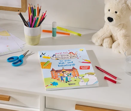 Ein weißer Schreibtisch mit Bastelmaterialien: ein Stapel Kinderzeichnungen, eine blaue Schere, ein Becher mit Buntstiften, Klebestift und Filzstifte. In der Mitte liegt ein Buch mit dem Titel „Margit Auer Die Schule der magischen Tiere Der große Mal- und Rätselspaß“. Rechts sitzen ein cremefarbener Teddybär, ein Glas Wasser und ein roter Filzstift.