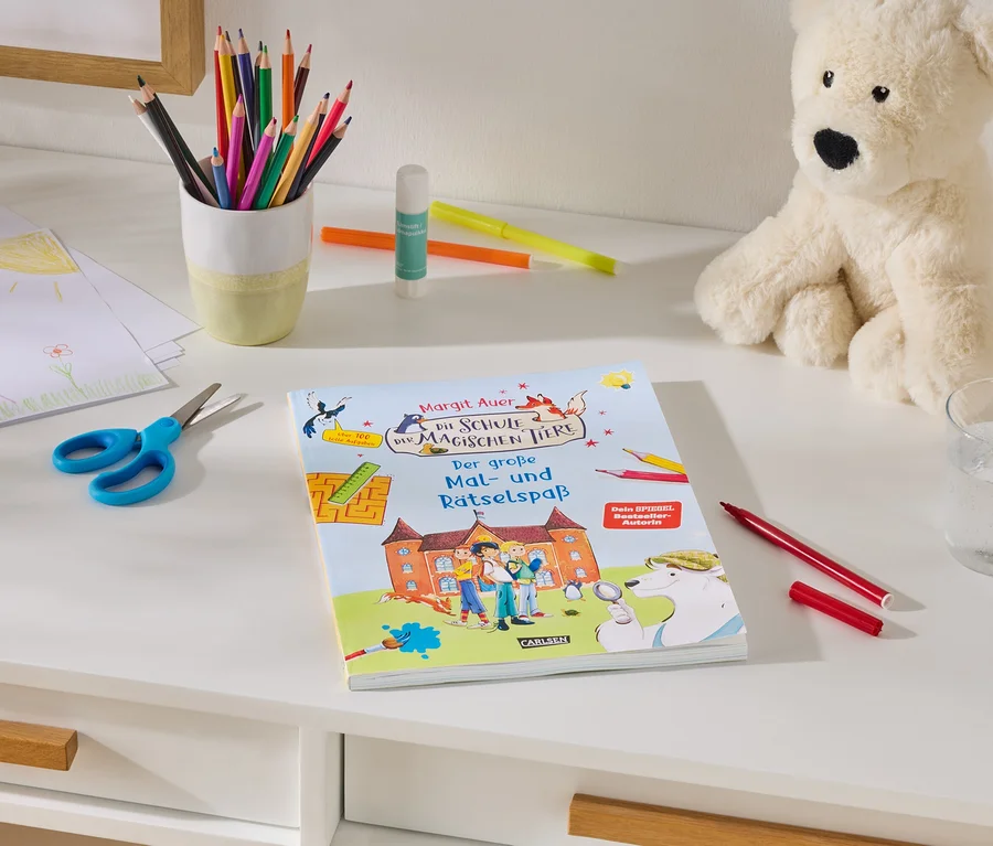 Ein weißer Schreibtisch mit Bastelmaterialien: ein Stapel Kinderzeichnungen, eine blaue Schere, ein Becher mit Buntstiften, Klebestift und Filzstifte. In der Mitte liegt ein Buch mit dem Titel „Margit Auer Die Schule der magischen Tiere Der große Mal- und Rätselspaß“. Rechts sitzen ein cremefarbener Teddybär, ein Glas Wasser und ein roter Filzstift.