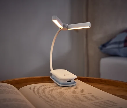 Eine weiße Klemm-Buchlampe beleuchtet ein offenes Buch auf einem Holztablett. Im Hintergrund ist ein Bett mit einem karierten Kissen zu sehen.