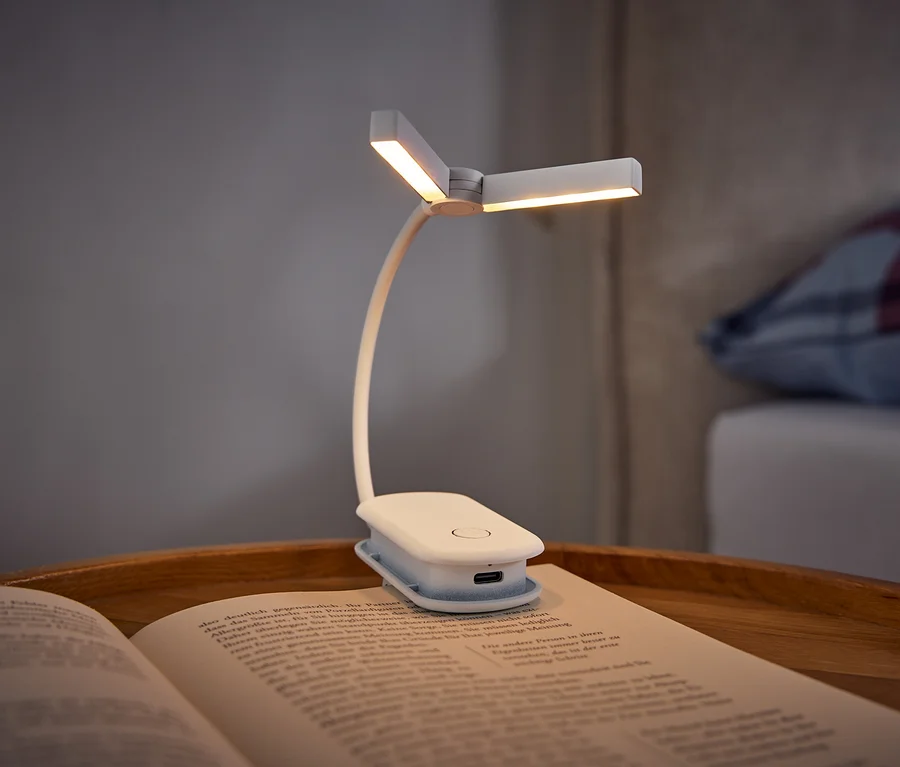 Eine weiße Klemm-Buchlampe beleuchtet ein offenes Buch auf einem Holztablett. Im Hintergrund ist ein Bett mit einem karierten Kissen zu sehen.