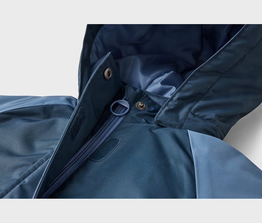 Nahaufnahme einer blauen Jacke, die den teilweise geöffneten Reißverschluss, eine Kapuze, Druckknöpfe und Klettverschlüsse an der Sturmklappe zeigt.