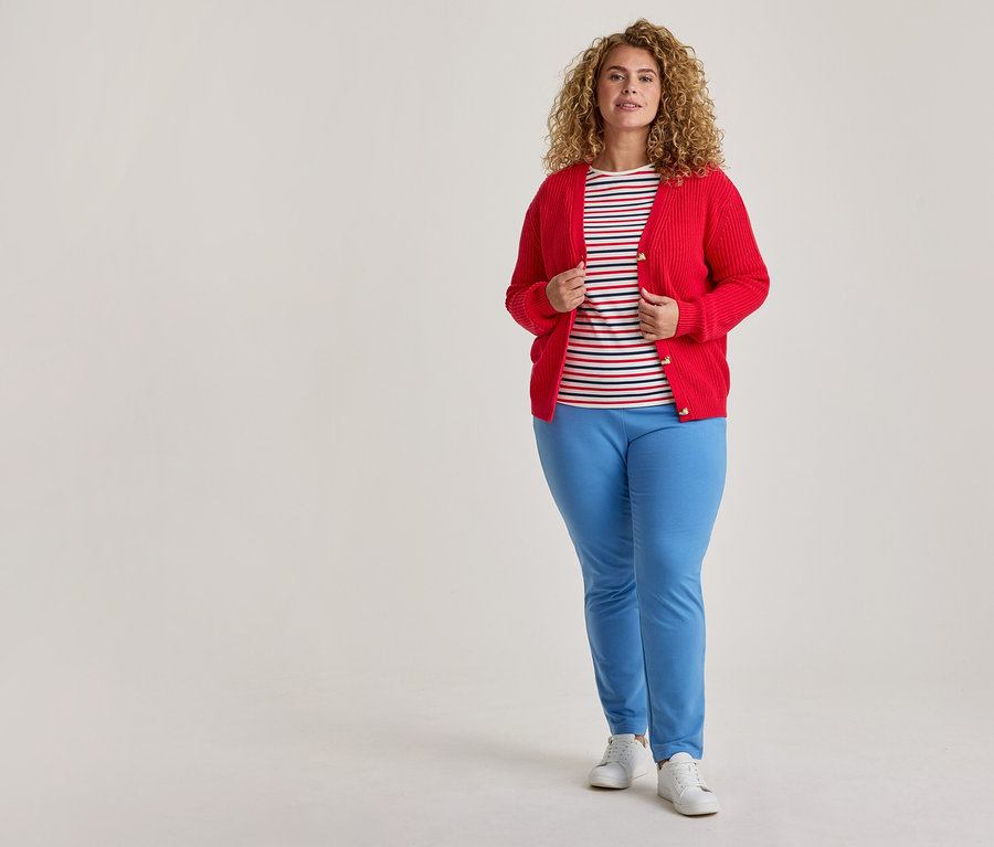 Eine Frau mit lockigem Haar steht und trägt einen offenen roten Cardigan über einem gestreiften T-Shirt, hellblauen Hosen und weißen Turnschuhen.