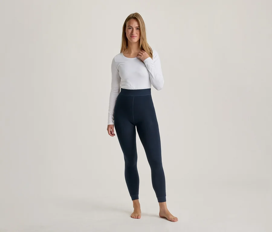 Eine Frau mit langen, hellbraunen Haaren steht barfuß, trägt ein weißes Langarmshirt und dunkelblaue Leggings und blickt nach vorne.