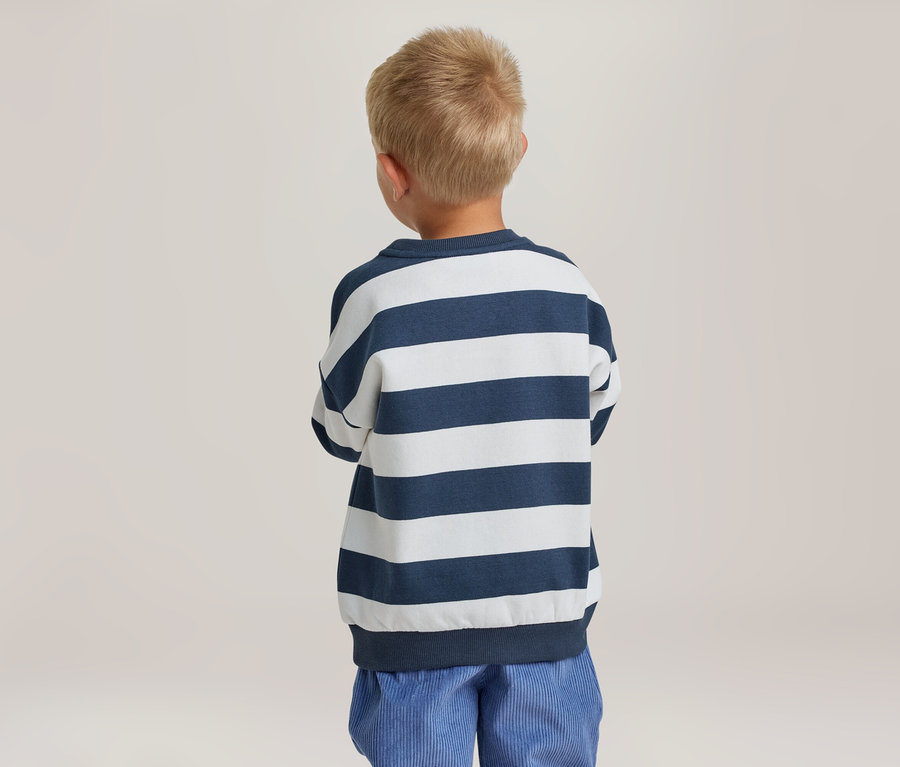 Kleiner Junge mit kurzen blonden Haaren steht mit dem Rücken zum Betrachter und trägt ein blau-weiß gestreiftes Langarmshirt und eine blaue Hose.