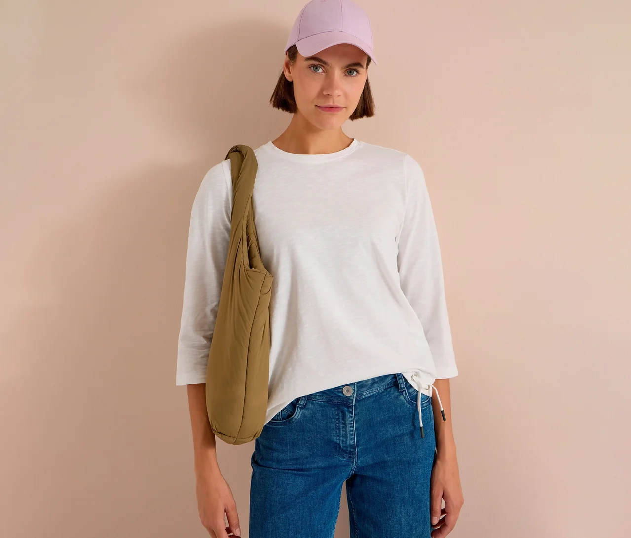 Frau mit hellvioletter Kappe, weißem CECIL 3/4-Shirt, blauen Jeans und khakifarbener, gepolsterter Umhängetasche.
