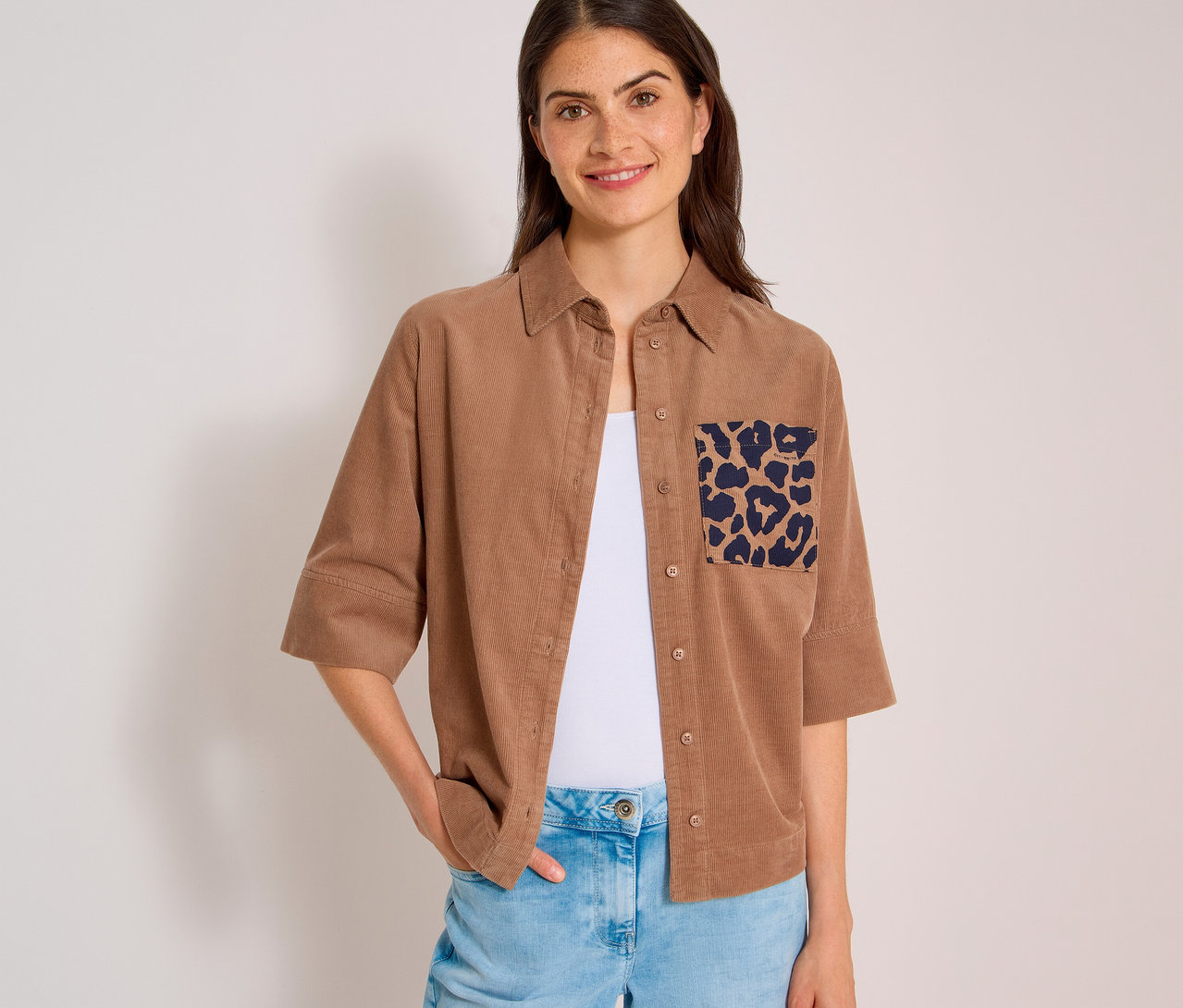 Lächelnde Frau in einer hellbraunen CECIL Hemdbluse aus Cord mit Leopardenmuster-Tasche über weißem Top und Jeans.