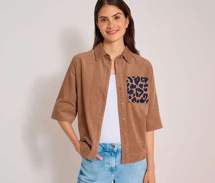 Lächelnde Frau in einer hellbraunen CECIL Hemdbluse aus Cord mit Leopardenmuster-Tasche über weißem Top und Jeans.