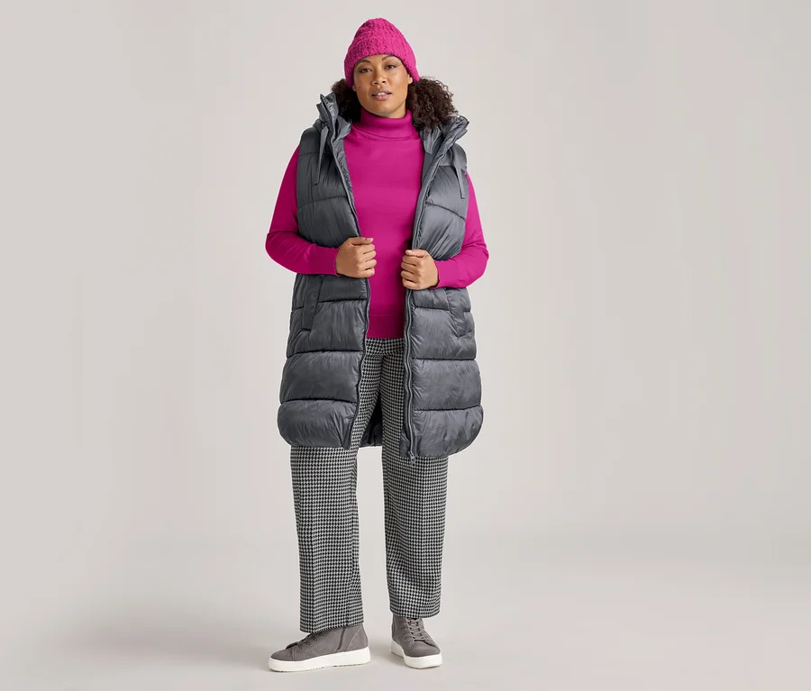 Frau mit pinker Mütze, pinkem Rollkragenpullover, schwarz-weiß karierter Hose und grauen Sneakers, posiert in einer langen anthrazitfarbenen Steppweste mit Kapuze.