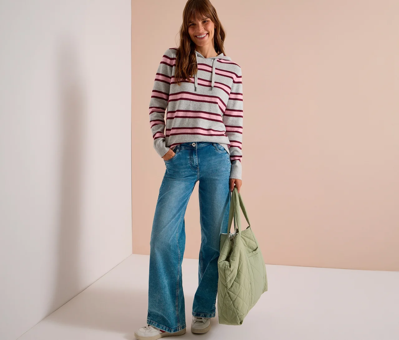 Eine lächelnde Frau steht mit den Händen in den Taschen und trägt einen grauen CECIL Strick-Hoodie mit rosa und dunkelroten Streifen, blaue Wide-Leg-Jeans und weiße Sneaker, während sie eine hellgrüne gesteppte Tragetasche hält.